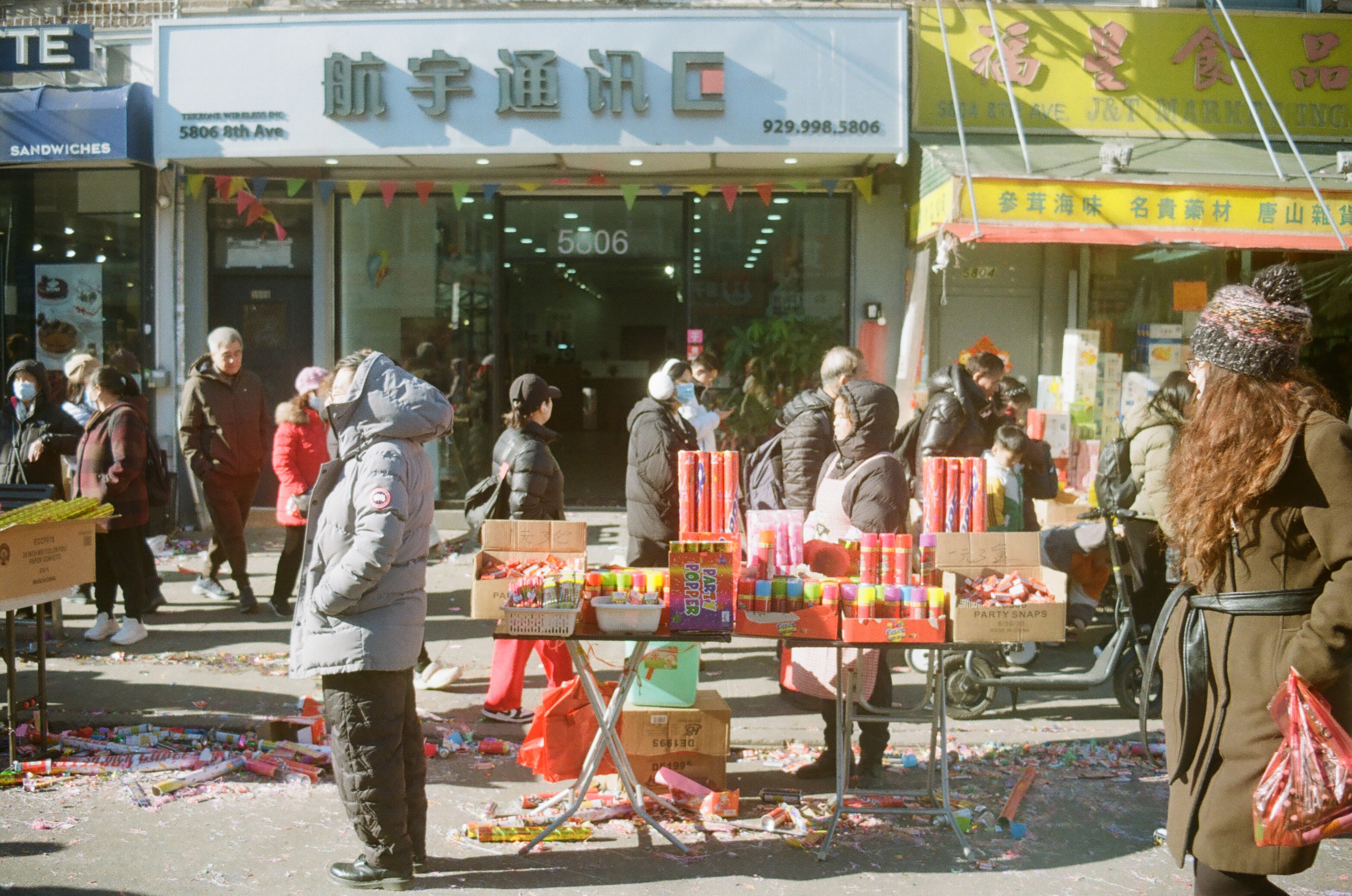 a view of a fireworks selling stand on a street littered with spent fireworks and celebratory confetti and other stuff, people standing around looking to the left